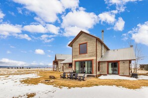 A two-story wooden cabin with a large sliding glass door, a deck with outdoor seating, and a clear view of the surrounding landscape. - Beautiful Targhee Farmhouse - Amazing Teton Views (Driggs)
