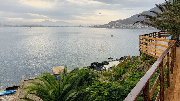 A coastal view with a wooden deck overlooking the sea, a small boat docked, and a distant mountain range. - SECHIN HOUSE ,  ubicada en el Balneario de Tortugas Caleta Norte (Tortuga Casma)