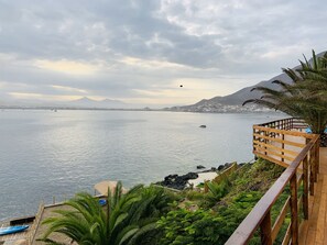 A coastal view with a wooden deck overlooking the sea, a small boat docked, and a distant mountain range. - SECHIN HOUSE ,  ubicada en el Balneario de Tortugas Caleta Norte (Tortuga Casma)