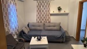 A small, cozy living room with a grey sofa, a white coffee table, and a small plant on a shelf.