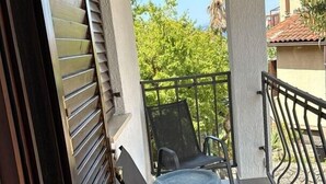A balcony with a chair and a table, a window with blinds, and a view of greenery and buildings. - Holiday apartment with balcony (Šilo)