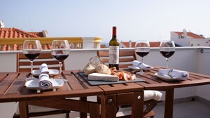 A wooden table set with wine glasses, a bottle of wine, bread, and cheese on a rooftop terrace with a view of buildings. - Ana's Apartments, Sea View (Sines)