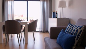 A modern living room with a sofa, a dining table, and chairs. There is a lamp on a side table and a view of the outdoors through large windows. - Ana's Apartments, Sea View (Sines)