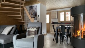 A modern living room with a grey sofa, a wooden staircase, a dining area with a black table and chairs, and a kitchen with wooden cabinets.