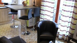 A living room with a black leather sofa, a glass coffee table, and two bar stools. There is a kitchen area with a bar counter and a dining table. - Holiday home in southern gardens with breathtaking panoramic views  (Canobio/Carmine)
