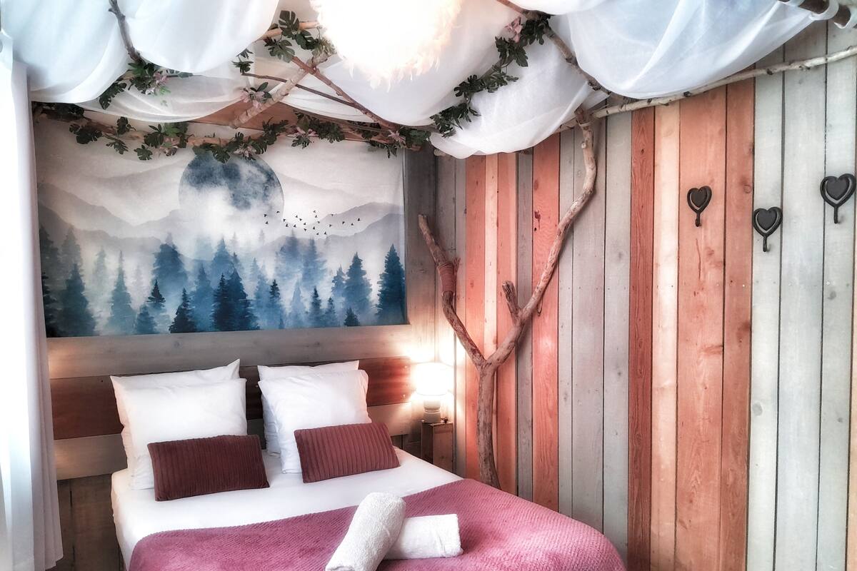 Une chambre à coucher avec un lit, un tableau représentant une forêt, une branche ornée de crochets en forme de cœur et un mur en bois.