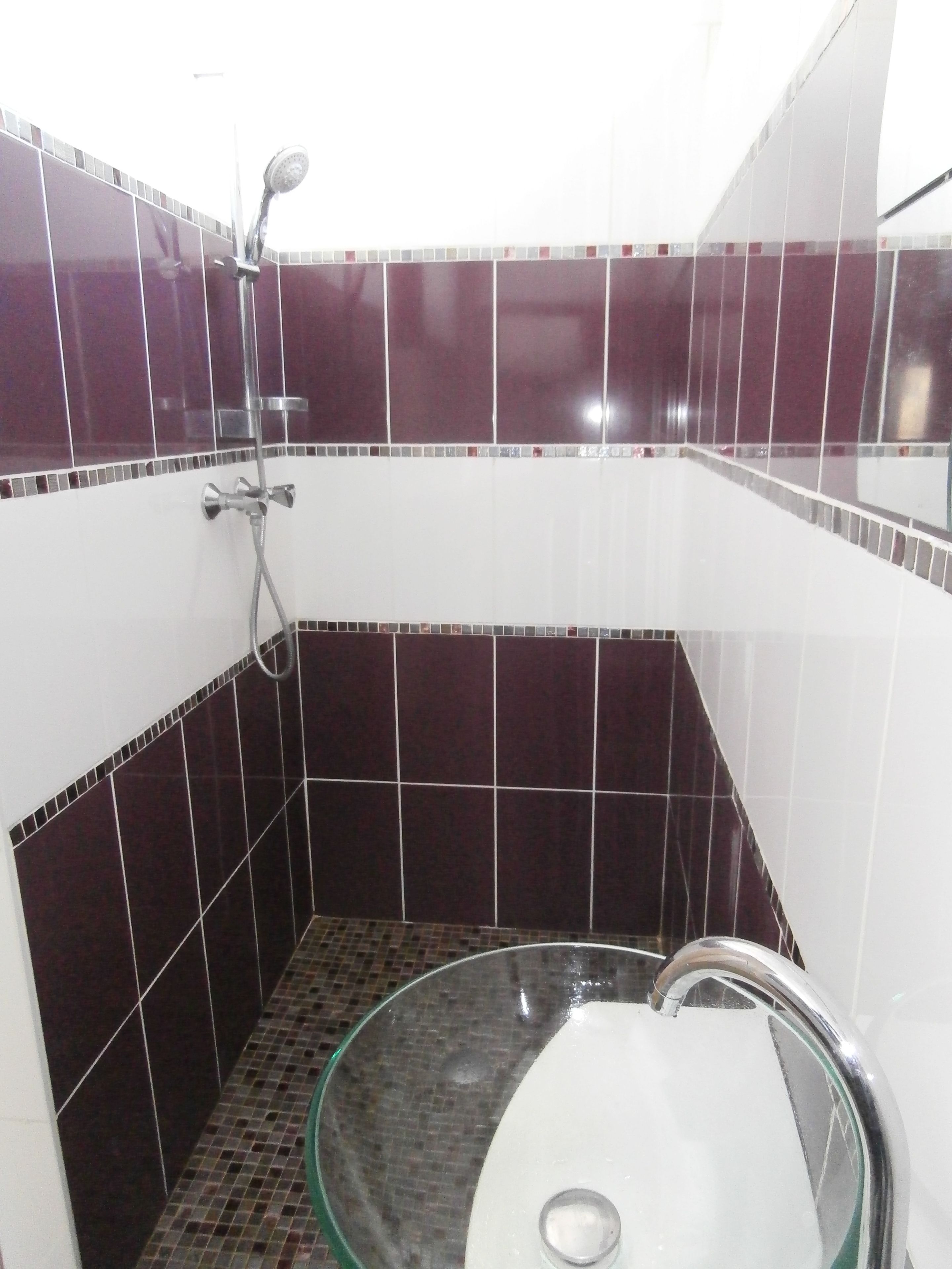 Un baño pequeño con un lavamanos de vidrio, una ducha y paredes cubiertas de azulejos rojo oscuro y blanco.