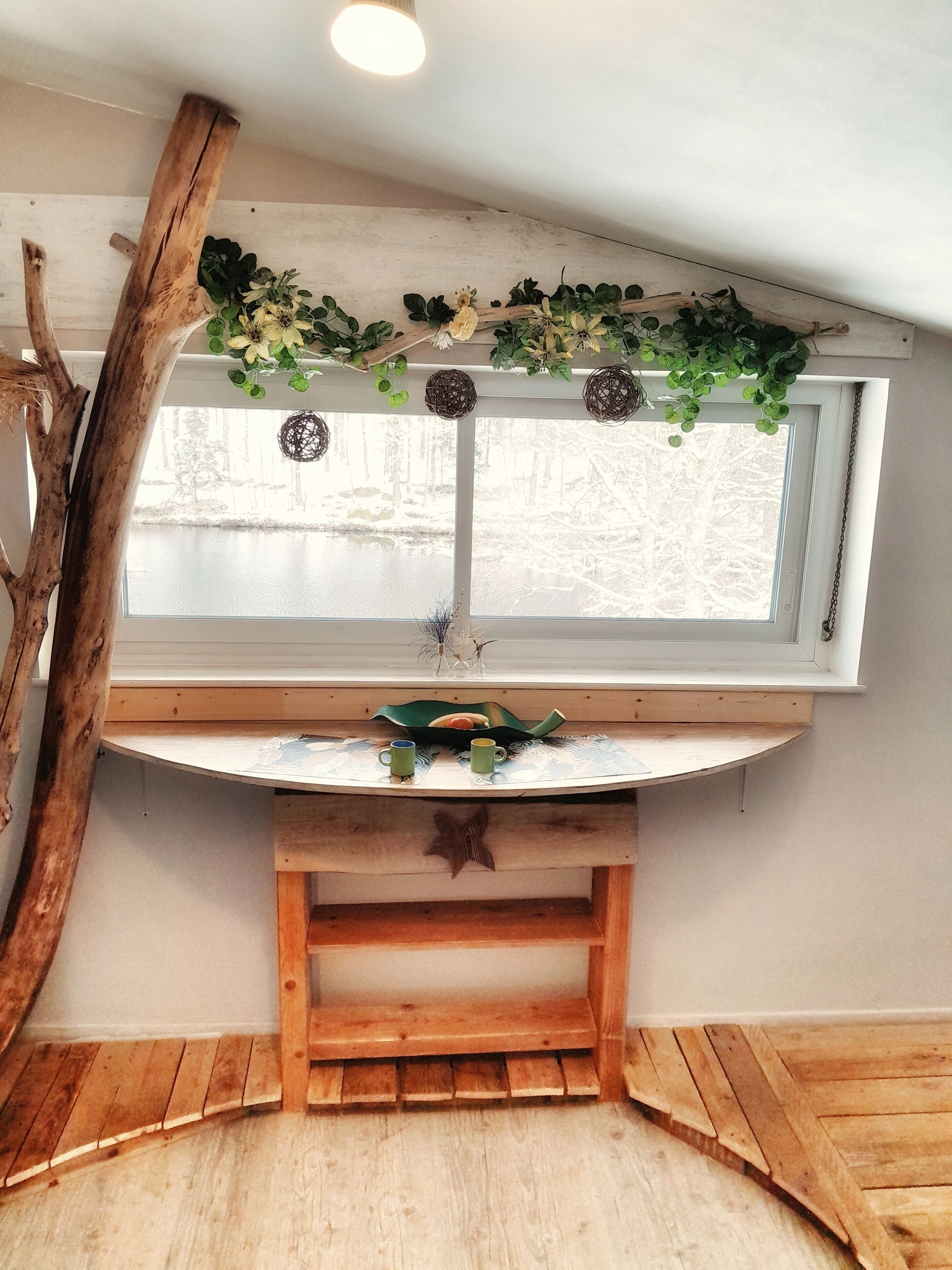 Une pièce rustique avec un banc en bois, une fenêtre donnant sur les arbres et une étagère ornée d’objets décoratifs.