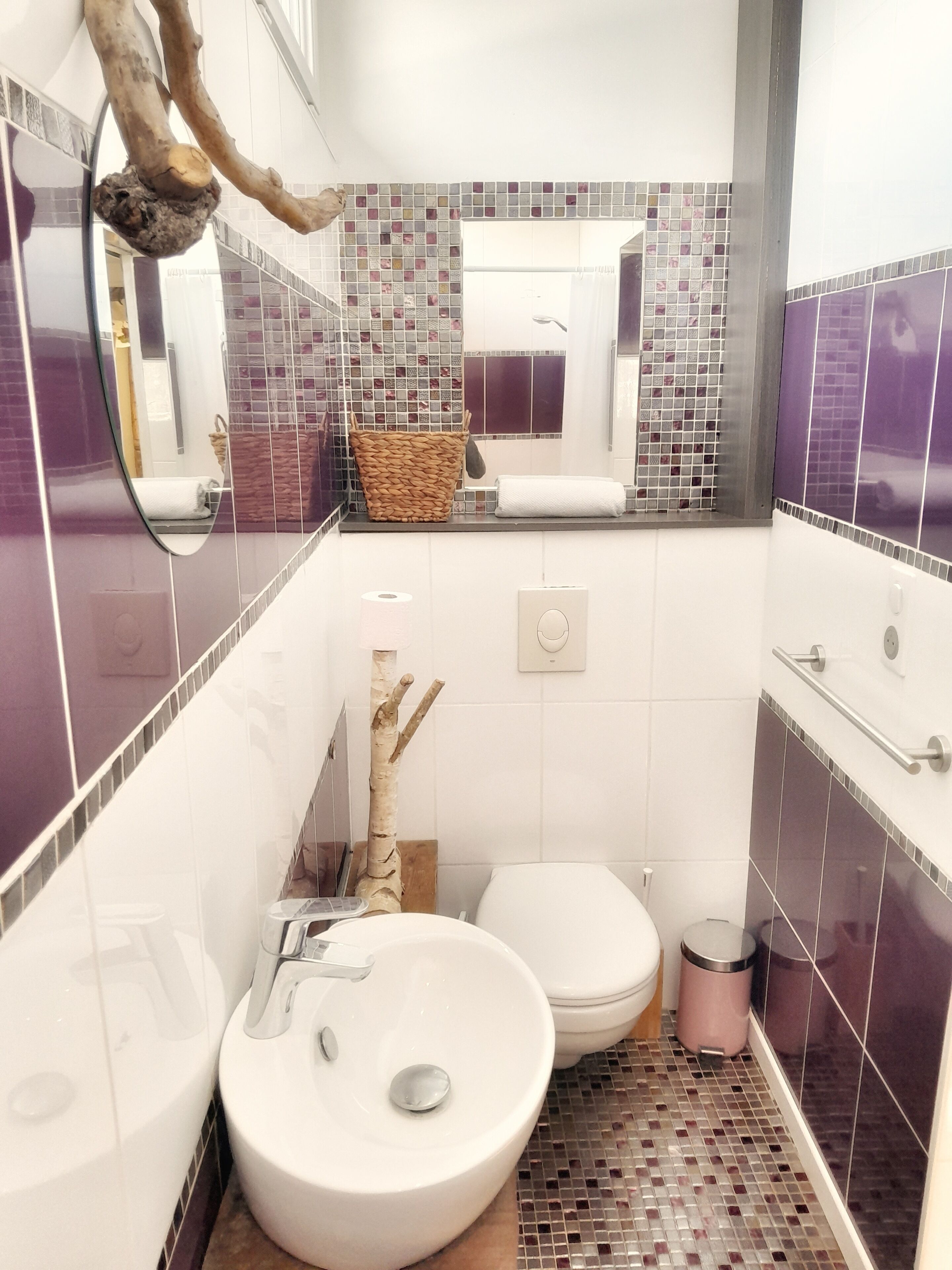 A small bathroom with a white sink, a toilet, and a mirror. There is a decorative branch and a woven basket on the counter.