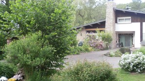 Une maison avec une cheminée en pierre, une véranda et un jardin bien entretenu, agrémenté de diverses plantes et fleurs.