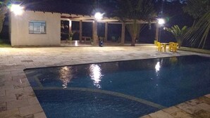 Ein Swimmingpool mit Fliesen umrandet, einem überdachten Sitzbereich und Außenbeleuchtung.