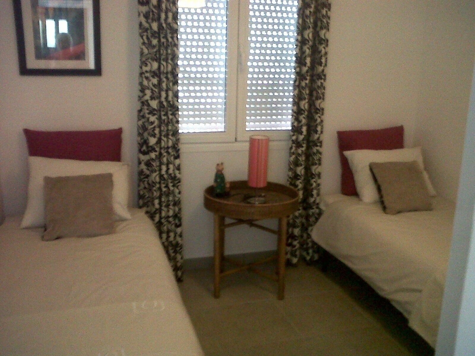 Una habitación pequeña y sencilla con dos camas, una mesita y una ventana con persianas.