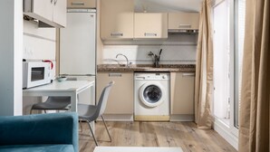 Een kleine keuken met een magnetron, wasmachine en een eethoek met een stoel.