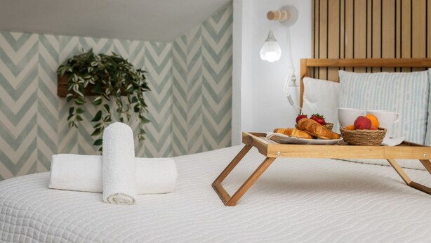 Een net opgemaakt bed met een witte deken, een opgerolde witte handdoek en een houten dienblad met croissants, fruit en een kop.