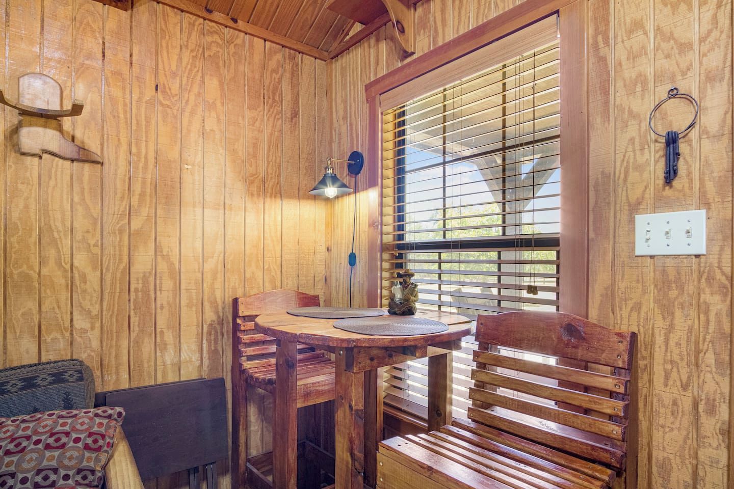 A wooden interior with a round table, two chairs, a bench, a wall-mounted light fixture, and a window with blinds.
