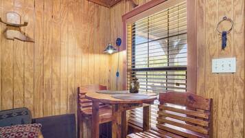 A wooden interior with a round table, two chairs, a bench, a wall-mounted light fixture, and a window with blinds.