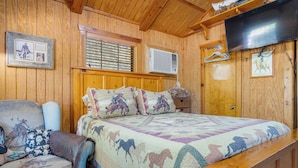 Ein Zimmer mit Holzwänden, einem Bett, einem Sessel, einem Fernseher und einem Fenster mit Jalousien.