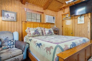 A wooden-paneled room with a bed, a chair, a television, and a window with blinds. - Double U Cowboy | 1BR Cabin with BBQ & Paddle Boat (Bandera)