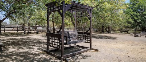 A wooden swing set in a park with a fenced area and trees.