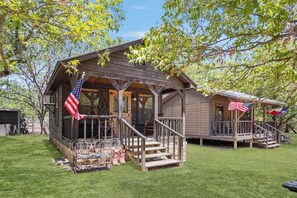 A wooden cabin with a porch, a covered deck, and two American flags. - Double U Cowboy | 1BR Cabin with BBQ & Paddle Boat (Bandera)