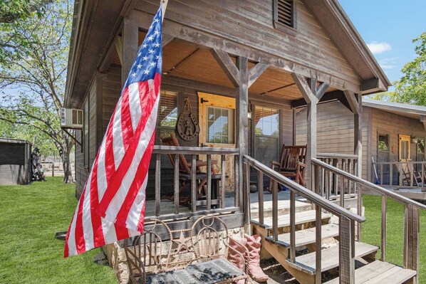A wooden house with a porch, a flag, and a welcome sign. - Double U Cowboy | 1BR Cabin with BBQ & Paddle Boat (Bandera)