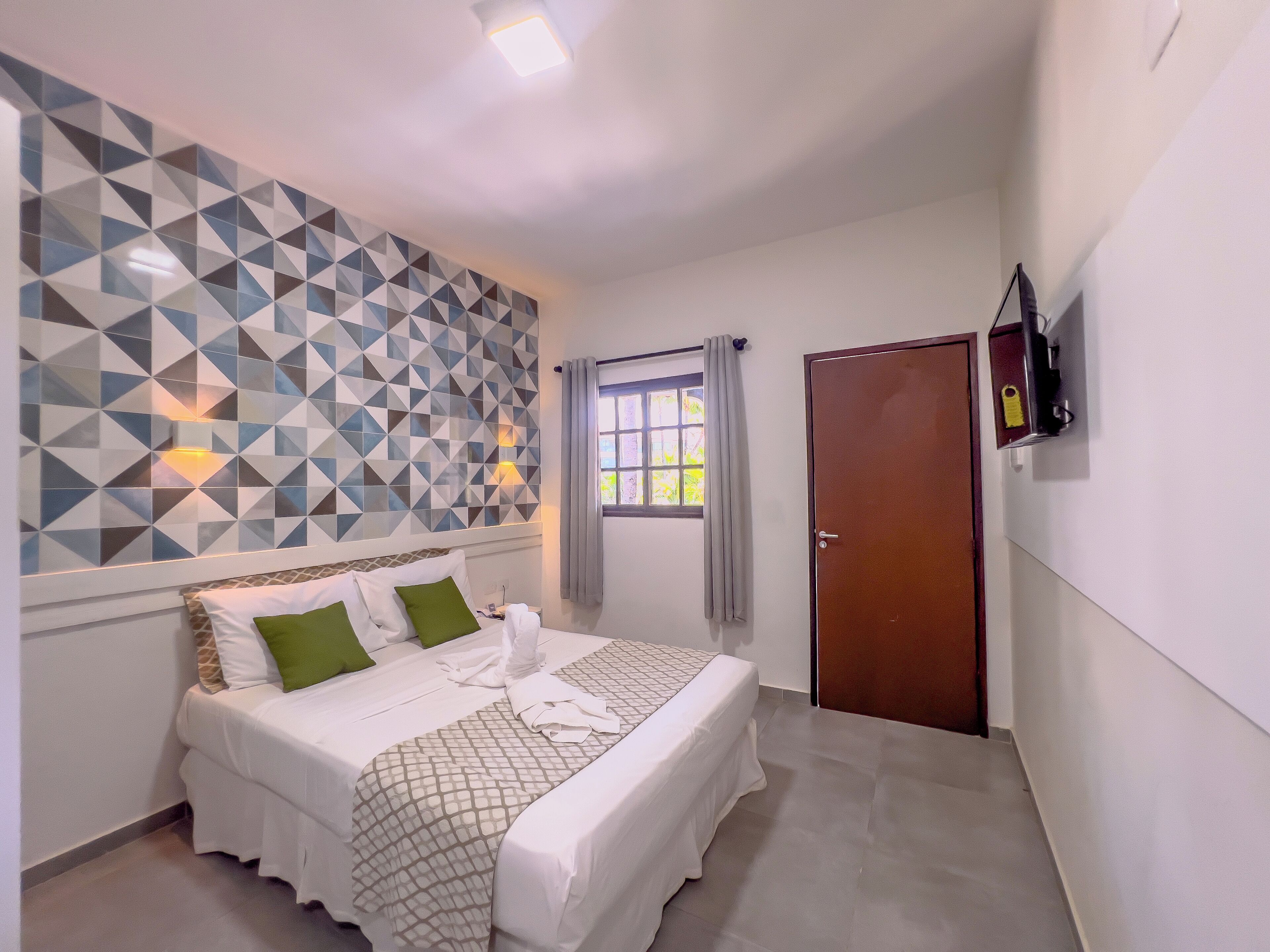 Un dormitorio con cama, pared con estampado geométrico, ventana con cortinas, puerta y televisor montado en la pared.