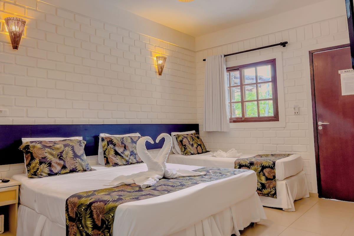 A hotel room with a white brick wall, two beds with floral bedding, a window with curtains, and wall-mounted lighting.