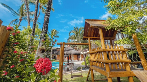 A wooden deck with a swing set, surrounded by lush greenery and a pool area. - Pousada Pérola do Porto (Ipojuca)