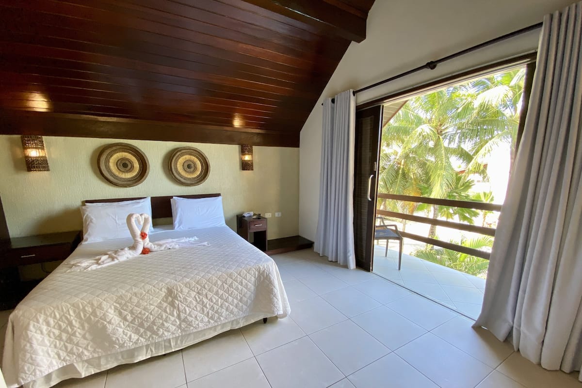 A hotel room with a large bed, a nightstand, a wall-mounted light fixture, and a view of palm trees through a sliding glass door.