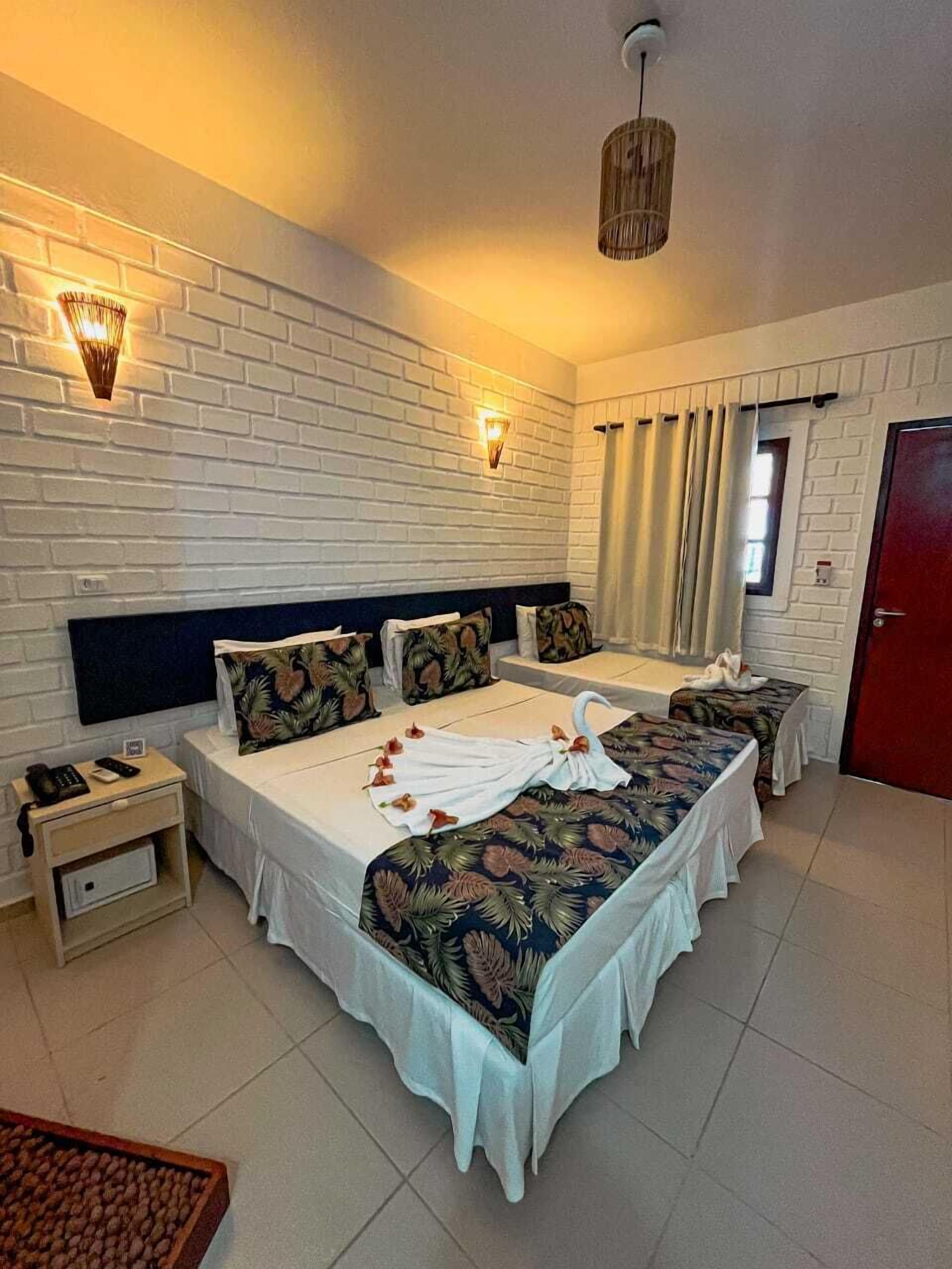 A hotel room with a large bed, two bedside tables, a nightstand, a red door, and a brick wall with two wall-mounted lights.