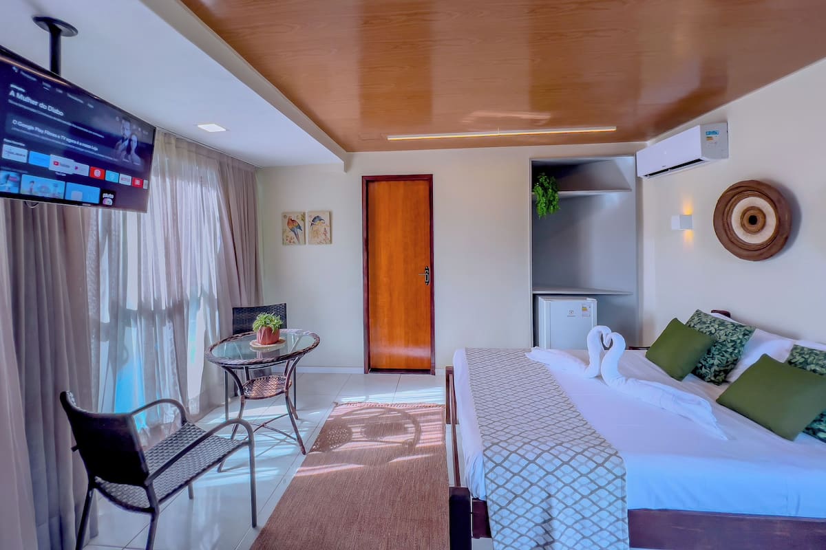 A hotel room with a bed, a desk with a chair, a television, a door, and a small table with a plant.