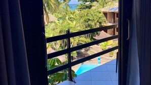 A view from a room with a sliding glass door to a balcony overlooking a pool, palm trees, and a building with a tiled roof. - Pousada Pérola do Porto (Ipojuca)