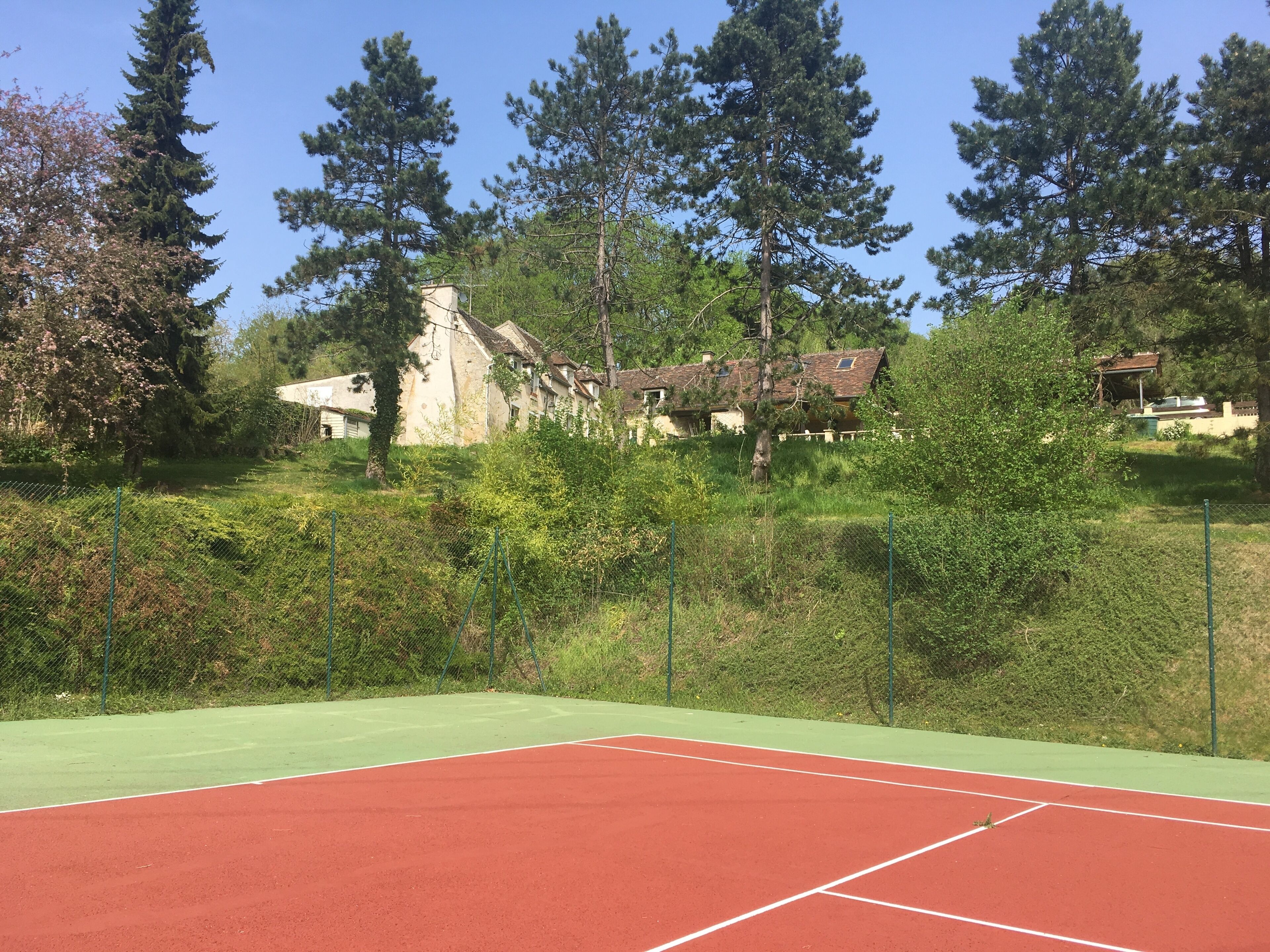 A tennis court with a green and red surface, surrounded by dense greenery and tall trees.
