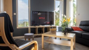 A modern living room with a large flat-screen TV displaying the Netflix logo, a black leather sofa, a wooden coffee table, and a view of greenery through large windows. - Ardèche Home - Gite le Nid - Comfortable house with swimming pool, rated 4*. (Tauriers)