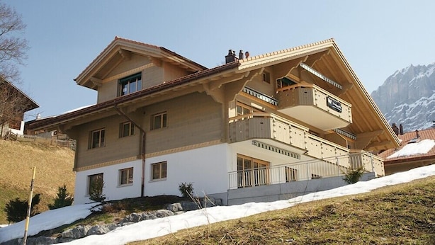 Een chalet met twee verdiepingen, een schuin dak, meerdere balkons en uitzicht op besneeuwde bergen.
