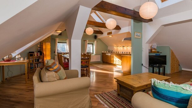 Ein geräumiger Loft mit hoher Decke, Holzbalken, einer gemütlichen Sitzecke mit Sofa und Sesseln, einem Esstisch und einer Küchenzeile.