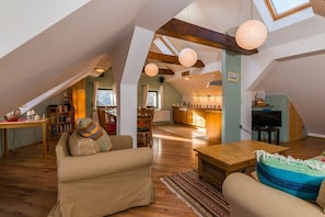 Ein geräumiger Loft mit hoher Decke, Holzbalken, einer gemütlichen Sitzecke mit Sofa und Sesseln, einem Esstisch und einer Küchenzeile.