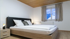 A bedroom with a wooden bed frame, white bedding, a nightstand with drawers, a bedside lamp, and a window with curtains.