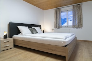 A bedroom with a wooden bed frame, white bedding, a nightstand with drawers, a bedside lamp, and a window with curtains.