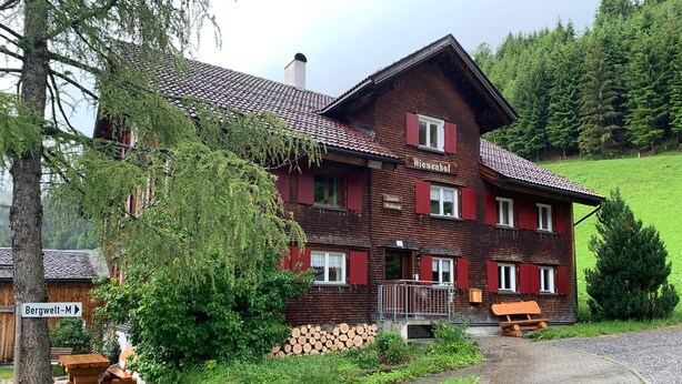 A traditional chalet-style building with a wooden facade, multiple windows with shutters, and a visible entrance with a wooden bench outside.