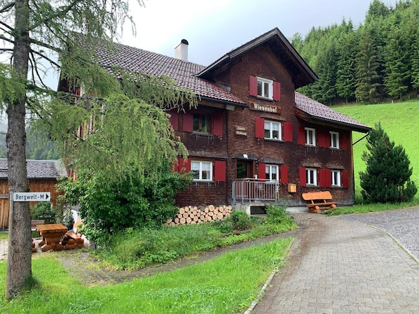 A traditional chalet-style building with a wooden facade, multiple windows with shutters, and a visible entrance with a wooden bench outside.