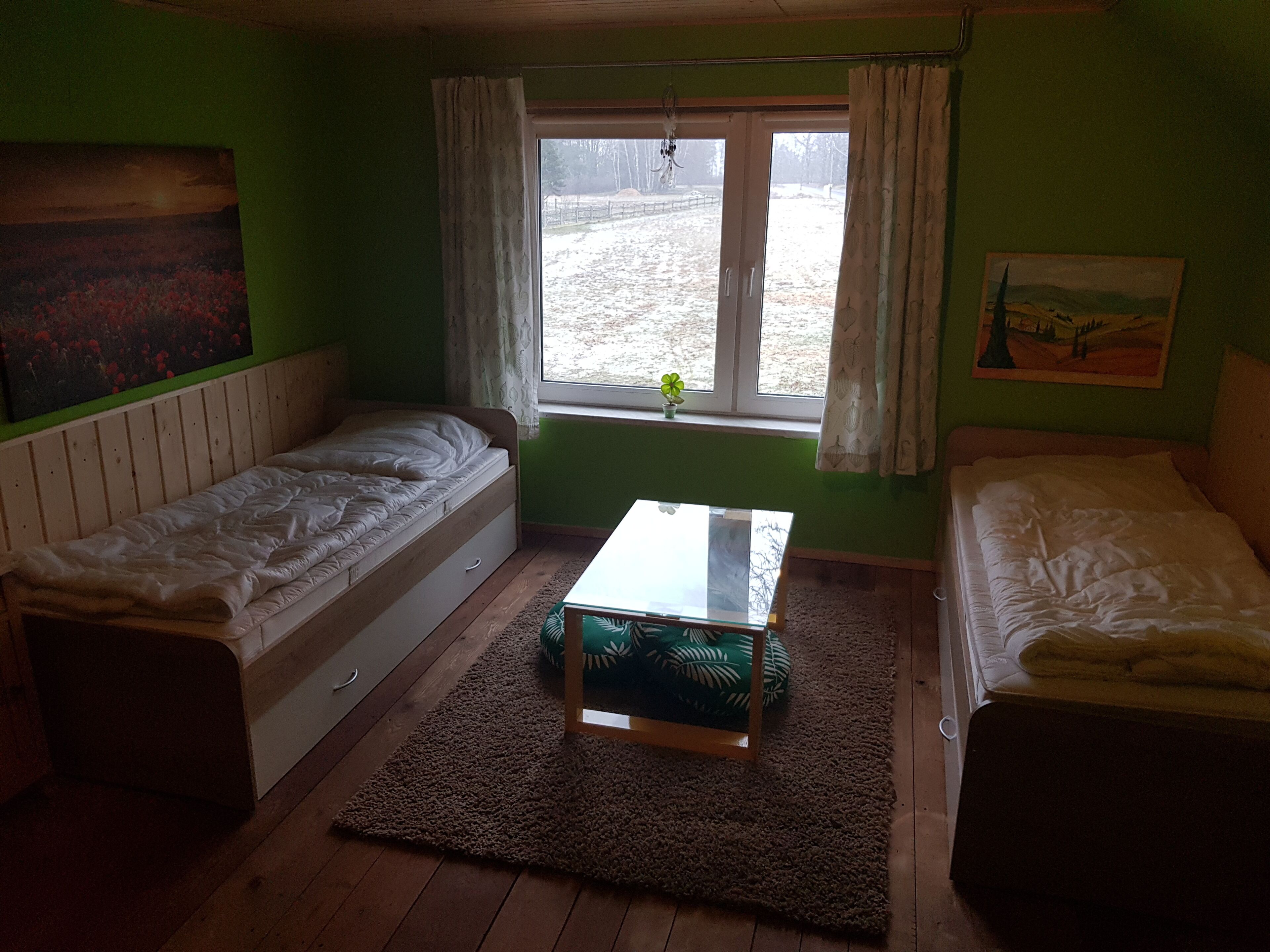 Une petite chambre accueillante, avec deux lits simples, un plancher en bois, une fenêtre avec des rideaux et un tableau au mur.