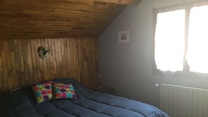 A bedroom with a wooden ceiling, a bed with a blue mattress and colorful pillows, a small bedside table, and a window with a view of the outside. - Cozy house in the countryside (Bolandoz)