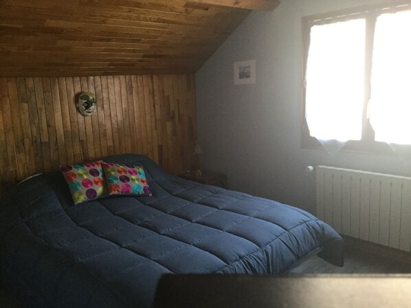 A bedroom with a wooden ceiling, a bed with a blue mattress and colorful pillows, a small bedside table, and a window with a view of the outside. - Cozy house in the countryside (Bolandoz)
