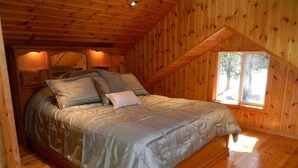 Une chambre confortable avec un plafond en bois et des murs inclinés, comprenant un grand lit avec une couette et des oreillers gris, une fenêtre donnant sur des arbres et un plancher en bois.