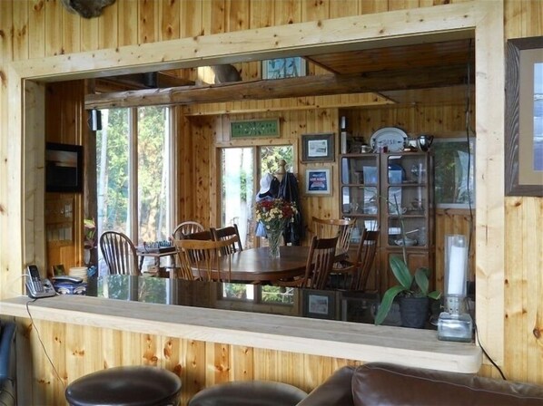 A wooden cabin interior with a dining area, wooden walls, and a large window overlooking greenery.