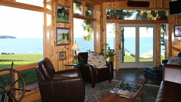Une cabane en bois dotée d’une grande fenêtre donnant sur une plage et l’océan. À l’intérieur, on trouve un canapé en cuir, une petite table avec une lampe et un jeu d’échecs.