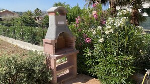 En udendørs stenovn i en have med fliseterrasse, omgivet af forskellige planter og blomster.