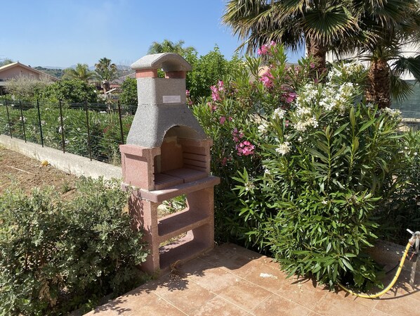 Forno de pedra ao ar livre em um jardim com piso de azulejos, rodeado por diversas plantas e flores.