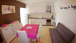 Une cuisine de taille modeste avec des meubles blancs, une table à manger recouverte d’une nappe rose et un canapé gris.